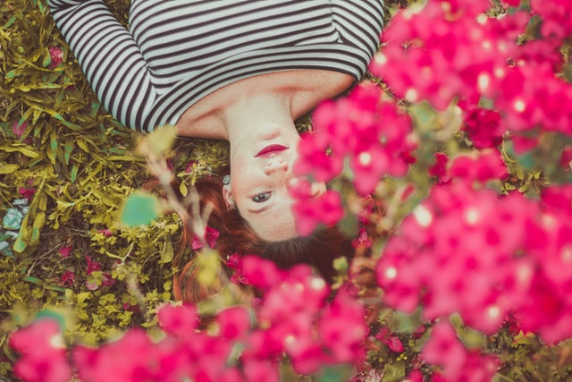 Uśmiechnięta kobieta leżąca na trawie, kadr przysłonięty różowymi kwiatami.