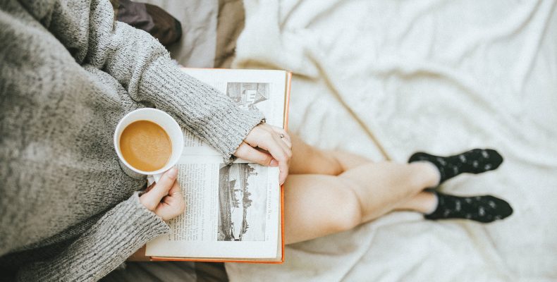 kobieta ubrana w szary sweter i czarne skarpetki z nogami opartymi o łóżko, czyta książkę i pije kawę, widok z góry