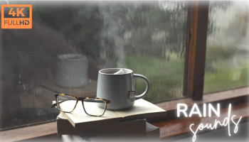 ksiązki, okulary i kubek z parującą herbatą na parapecie okna, za którym pada deszcz
