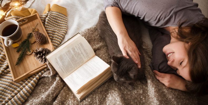 kobieta leżąca na łóżku z książką i kawą, głaskająca kota