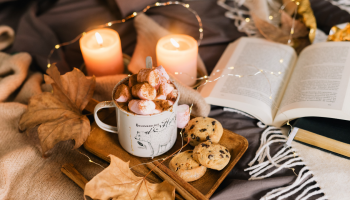 gorąca czekolda z piankami, ciastka oraz książka wśród zapalonych świec i światełek na łóżku