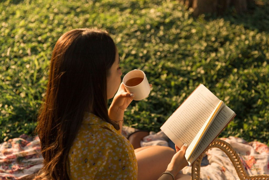 kobieta siedzi na kocu na trawie, w ręku trzyma kubek i książkę
