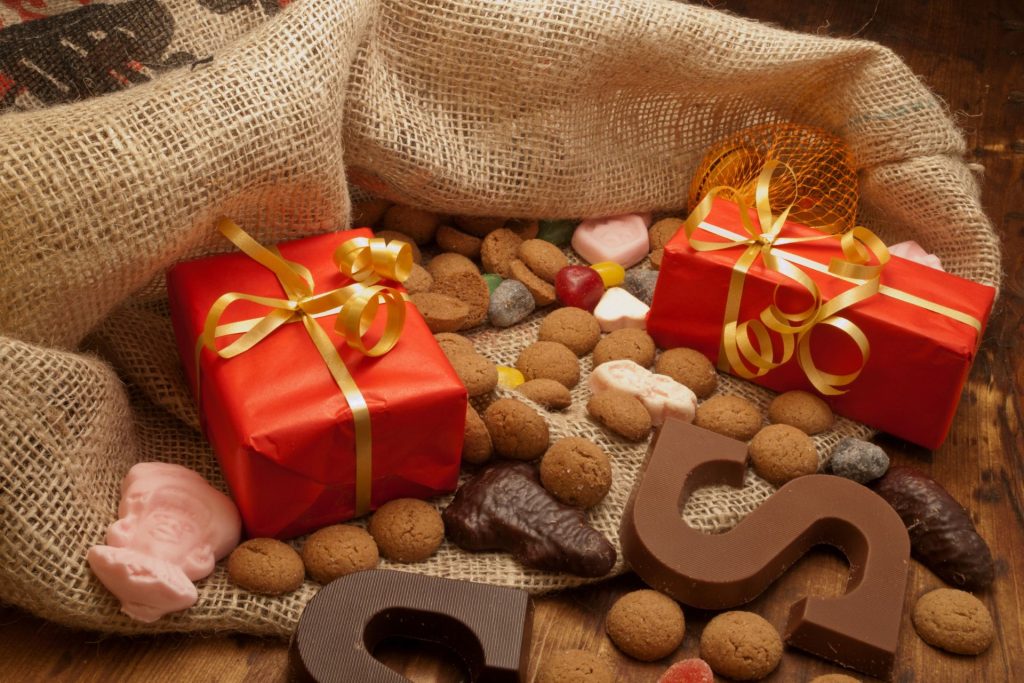 dwa prezenty zapakowane w czerwony papier ze złotymi kokardkami, rozsypane ciastka i czekoladki, zaraz obok lniany worek Mikołaja, wszystko leży na drewnianym stole