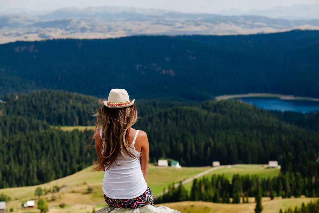 dziewczyna w kapeluszu siedzi na wzniesieniu i patrzy się na góry pokryte lasem i jezioro