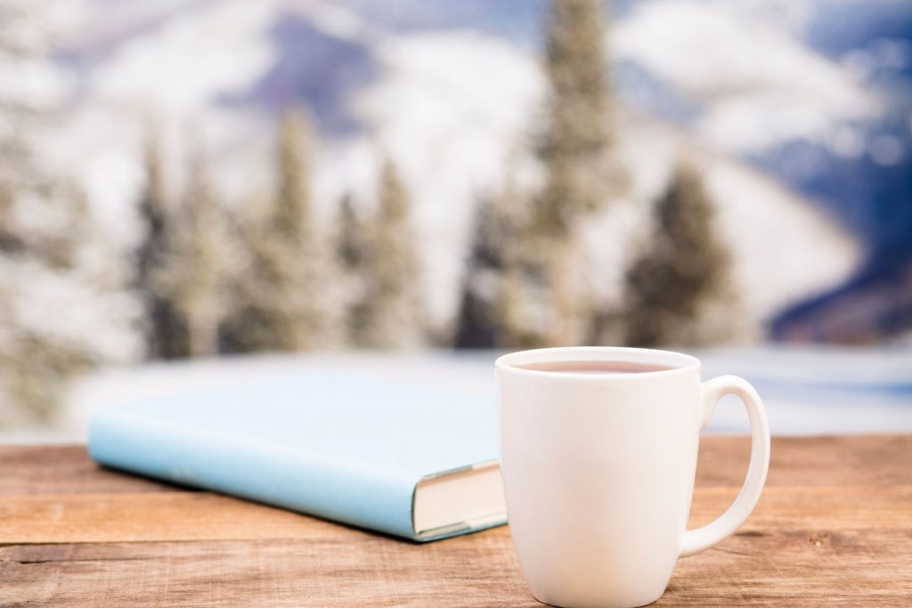książka z niebieską okładką i biały kubek stoi na drewnianym blacie, w tle widać ośnieżone choinki i zimową aurę