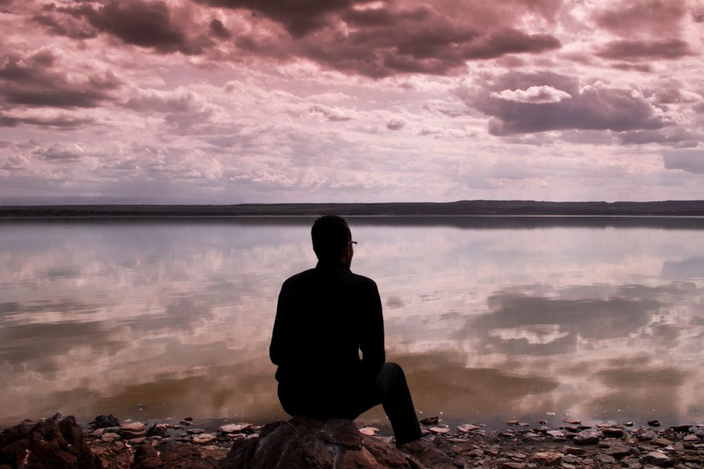 mężczyzna siedzi nad brzegiem jeziora i rozmyśla, plaża, różowe chmury na niebie